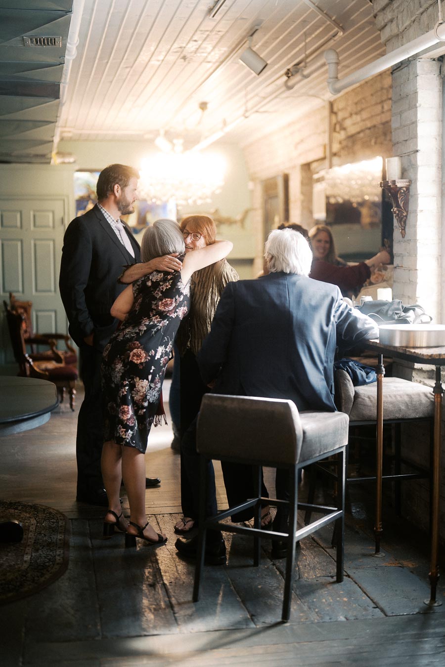 A group of people in a warmly lit, stylish room, embracing and socializing, creating a sense of warmth and connection. The setting includes vintage decor and a chandelier, suggesting a cozy and elegant gathering atmosphere.