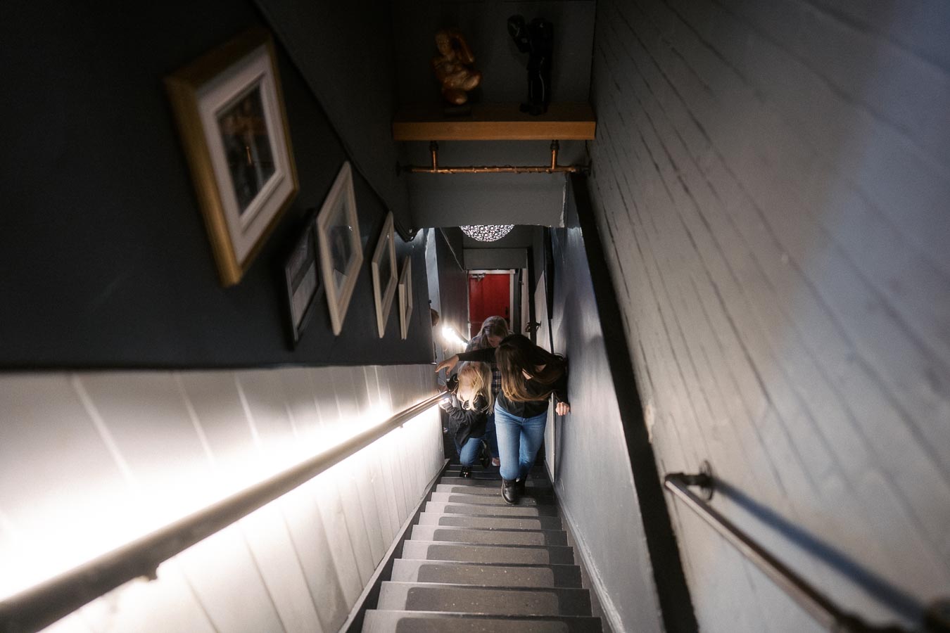 A dimly lit, narrow staircase with framed pictures on the wall and people descending, creating a sense of depth and perspective.