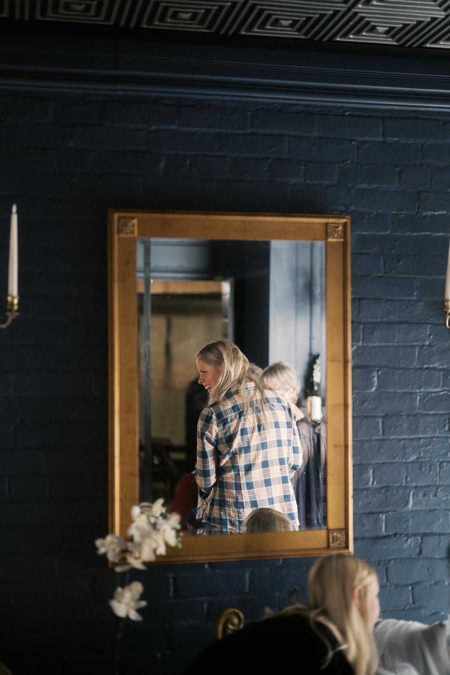 Mirror reflection of a smiling person in a plaid shirt against a dark blue brick wall, featuring elegant wall sconces and an ornate ceiling pattern.