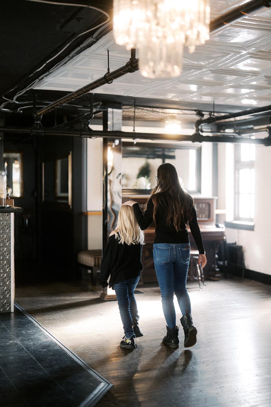 A woman and child walking hand in hand through a modern industrial-style room with a piano and chandelier.