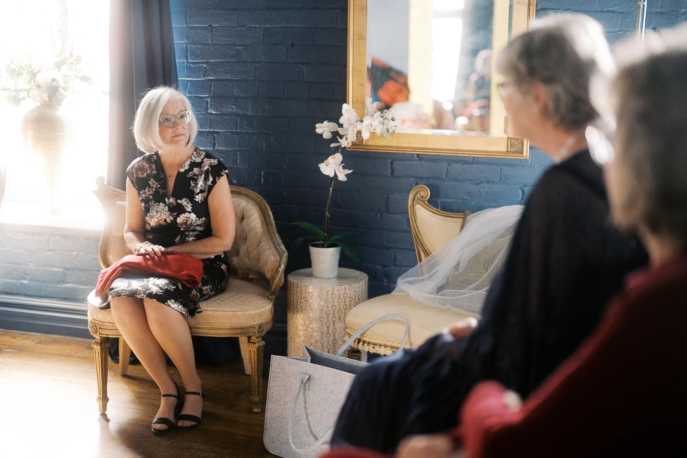 Elegant woman sitting on antique chair in stylish room with blue brick wall, floral dress, and natural light; reflection in mirror and decorative potted orchid visible.