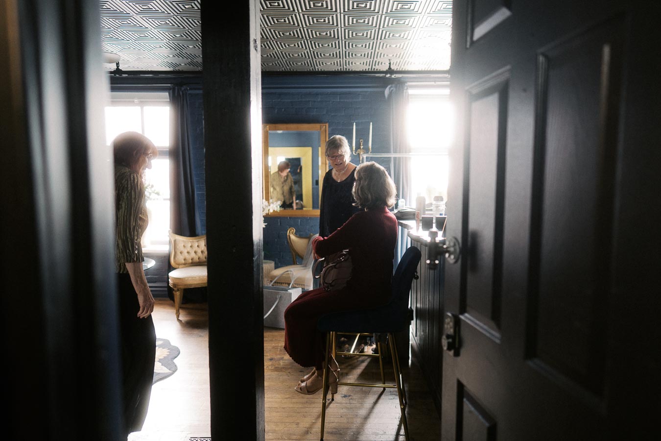 A group of women engaging in conversation in a stylish room with modern decor, featuring elegant furniture, a patterned ceiling, and natural light streaming through a window.