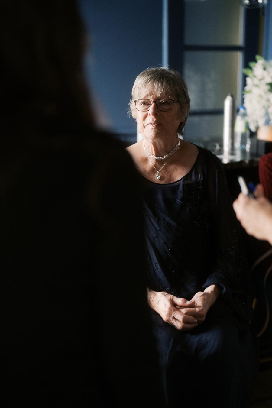Elderly woman in glasses and a dark dress sits pensively at a formal event, against a softly lit background with flowers and bottles.