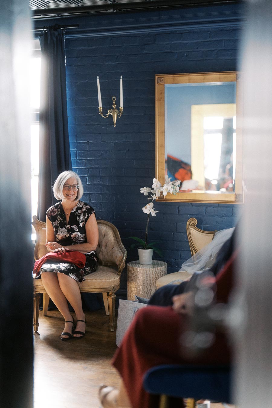 A woman with gray hair wearing a floral dress sits on a vintage chair in a stylish room with navy blue walls, decorated with a wall sconce and a framed mirror reflecting light from a window. An orchid plant sits on a side table nearby, adding elegance to the setting.