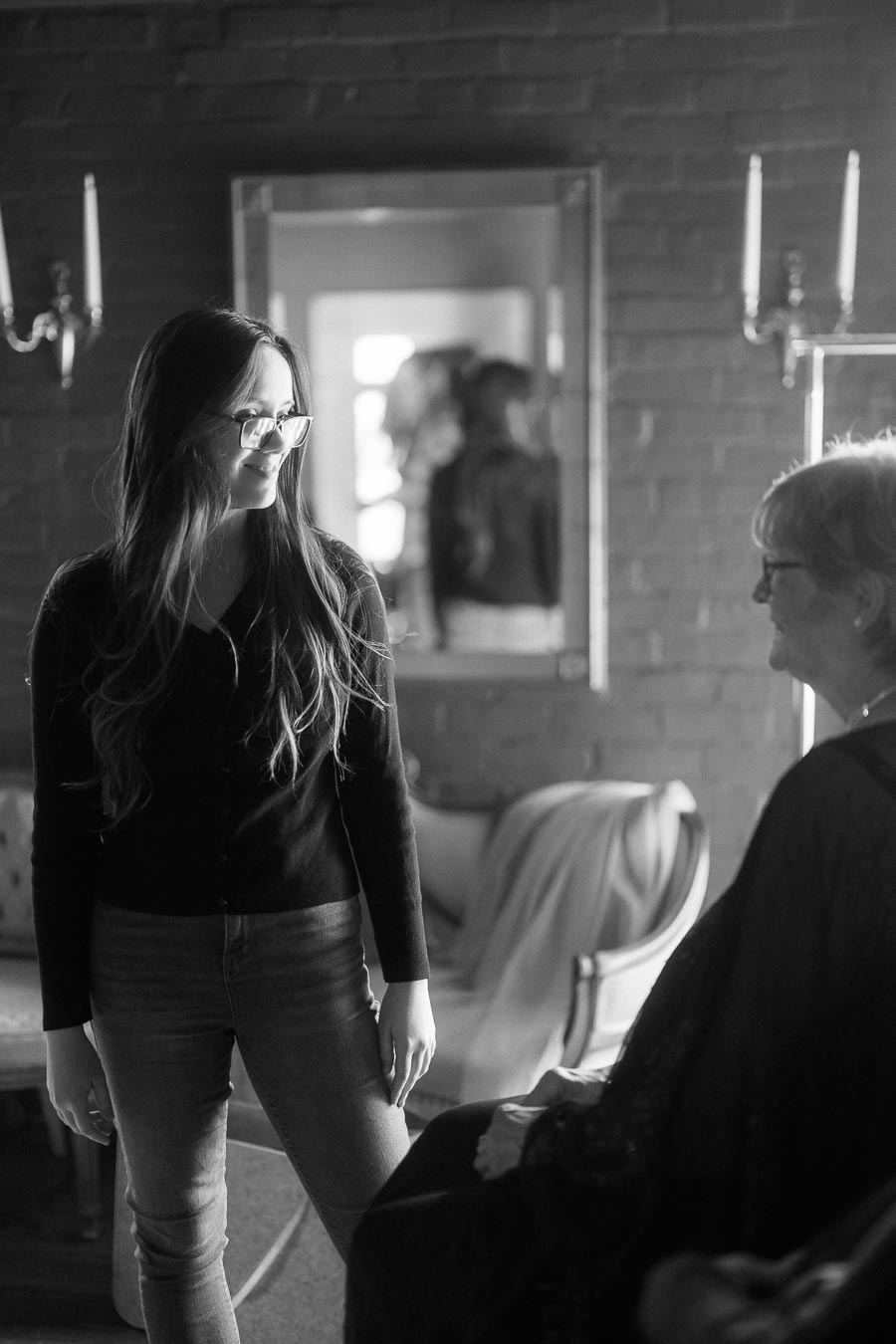 Black and white photo of two women smiling in a cozy room, one standing, wearing glasses and casual clothes, the other seated, engaging in conversation by a mirror with soft lighting.