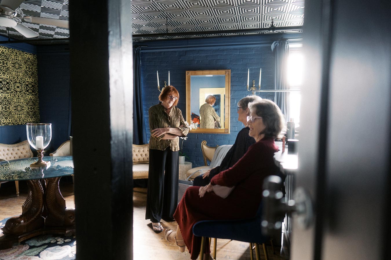 A group of people engaged in conversation in a stylish, modern living room with blue walls, a geometric patterned ceiling, and elegant furniture. A large mirror and decorative items add to the room’s sophisticated ambiance.