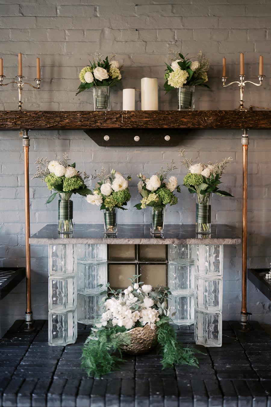 Elegant rustic floral decor featuring green and white bouquets in clear glass vases on a wooden shelf, complemented by candles and metal candlesticks, set against a textured brick wall.