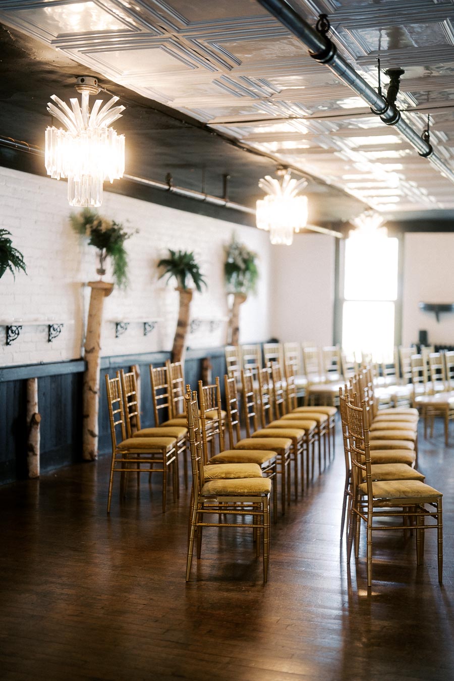 Elegant wedding ceremony setup with rows of gold Chiavari chairs, hardwood floor, and modern chandeliers, creating a sophisticated and stylish venue atmosphere.