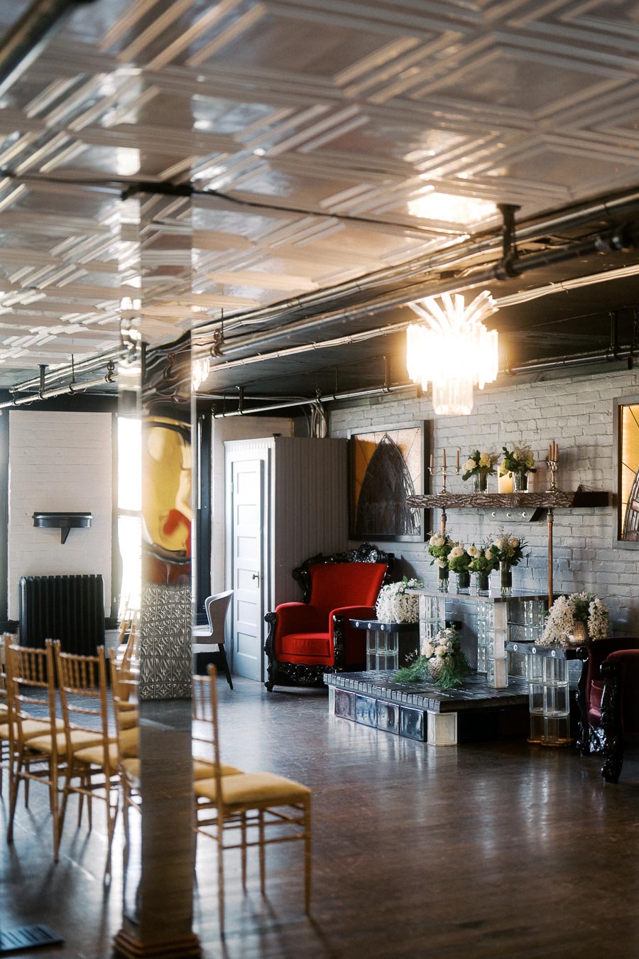 Elegant interior of a modern event space featuring a stylish red armchair, chic floral arrangements, and ambient lighting reflecting off a sleek tin ceiling.