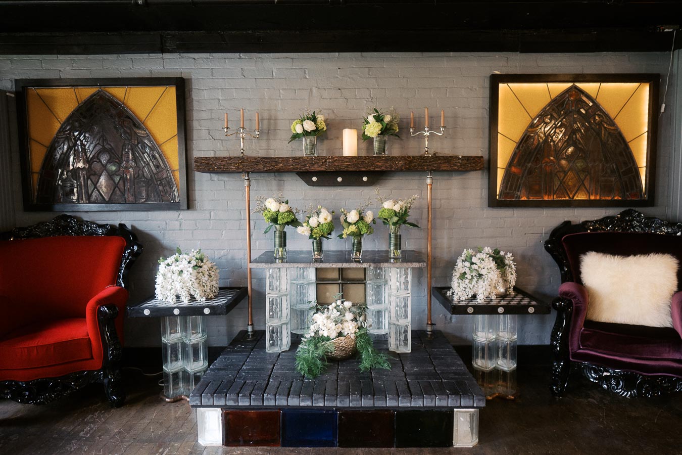 Cozy living room with vintage red and purple armchairs flanking a stylish fireplace. Decor includes stained glass artwork, a wooden mantle featuring elegant candelabras, and floral arrangements in glass vases, enhancing the room's warm ambiance.