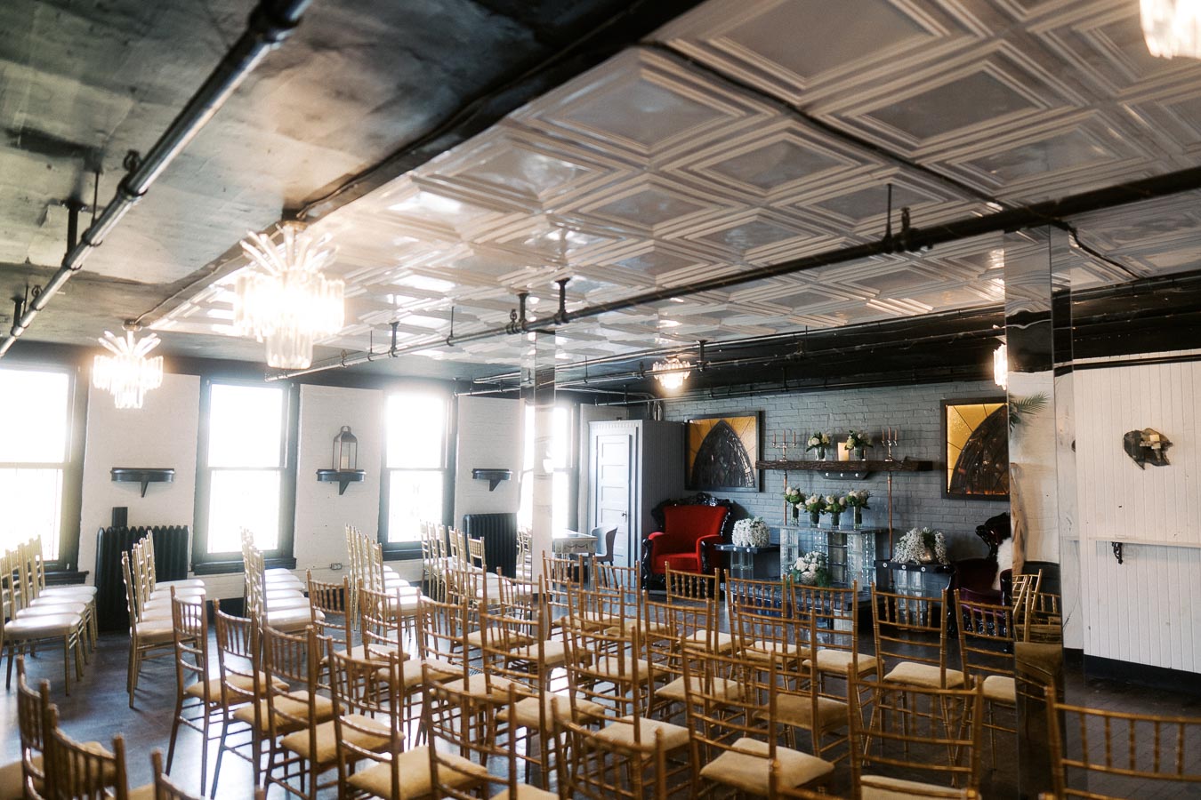 Chic industrial event space with elegant wooden chairs and modern chandeliers, featuring a stylish seating area adorned with floral arrangements and decorative artwork. Ideal for weddings and intimate gatherings.
