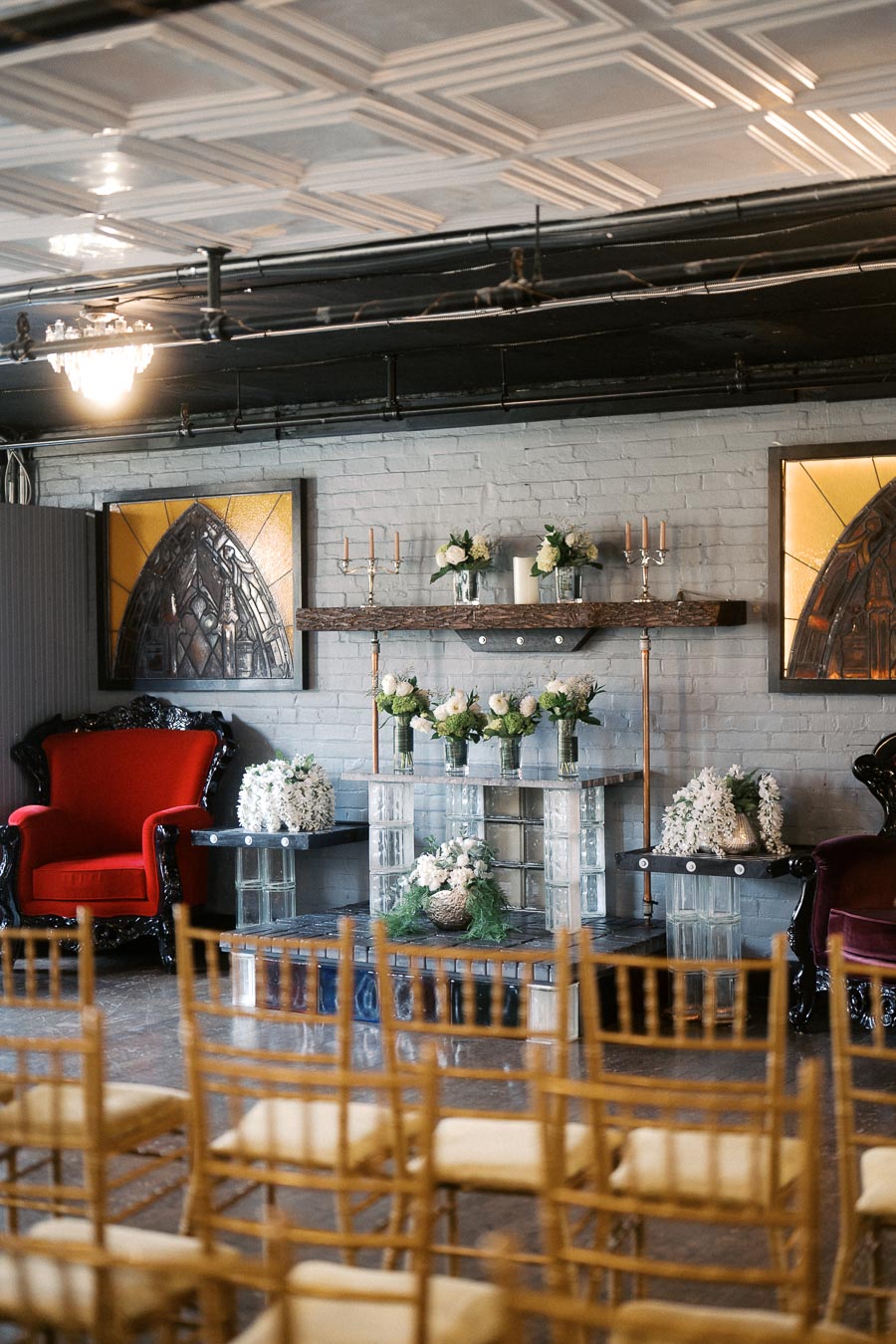 Elegant wedding venue interior with floral arrangements and vintage decor, featuring a red velvet armchair, glass brick altar with white flowers, and gold chairs arranged for a ceremony.
