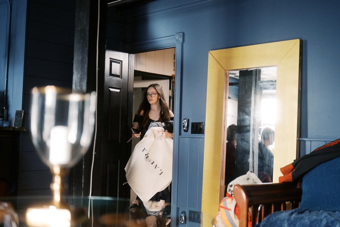 Woman entering room with blue walls and large yellow mirror, holding a garment bag and smartphone, with blurred foreground objects and cozy interior setting.