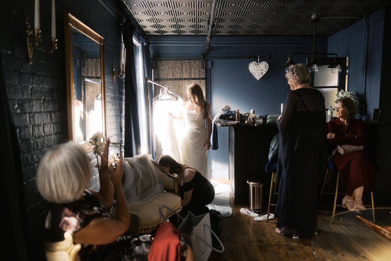 Bride in white dress preparing for wedding in dimly lit room surrounded by women, one taking photos, highlighting a cozy and intimate atmosphere.