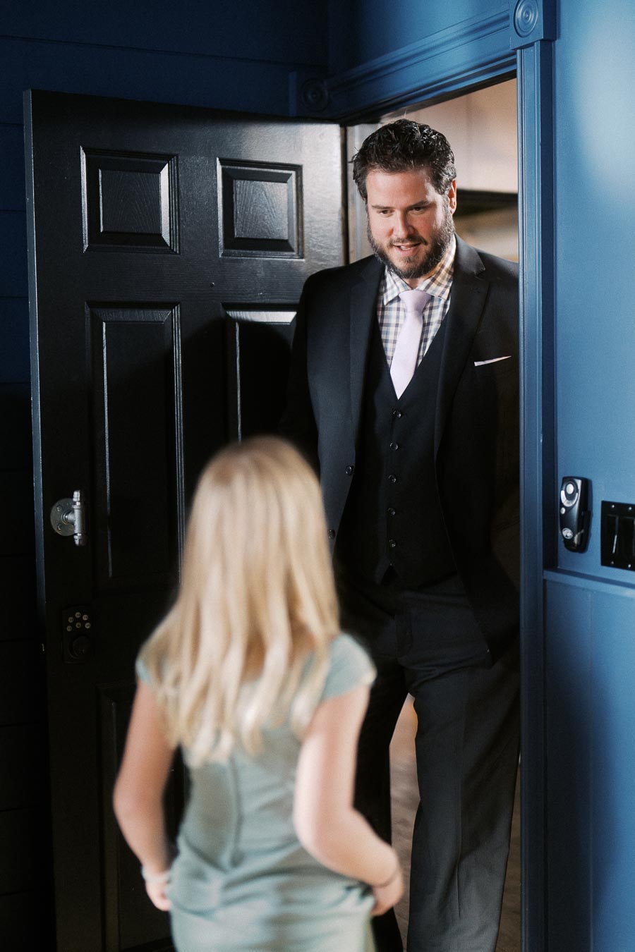 Man in a suit smiling at a child in a doorway, with blue walls and a black door in the background.