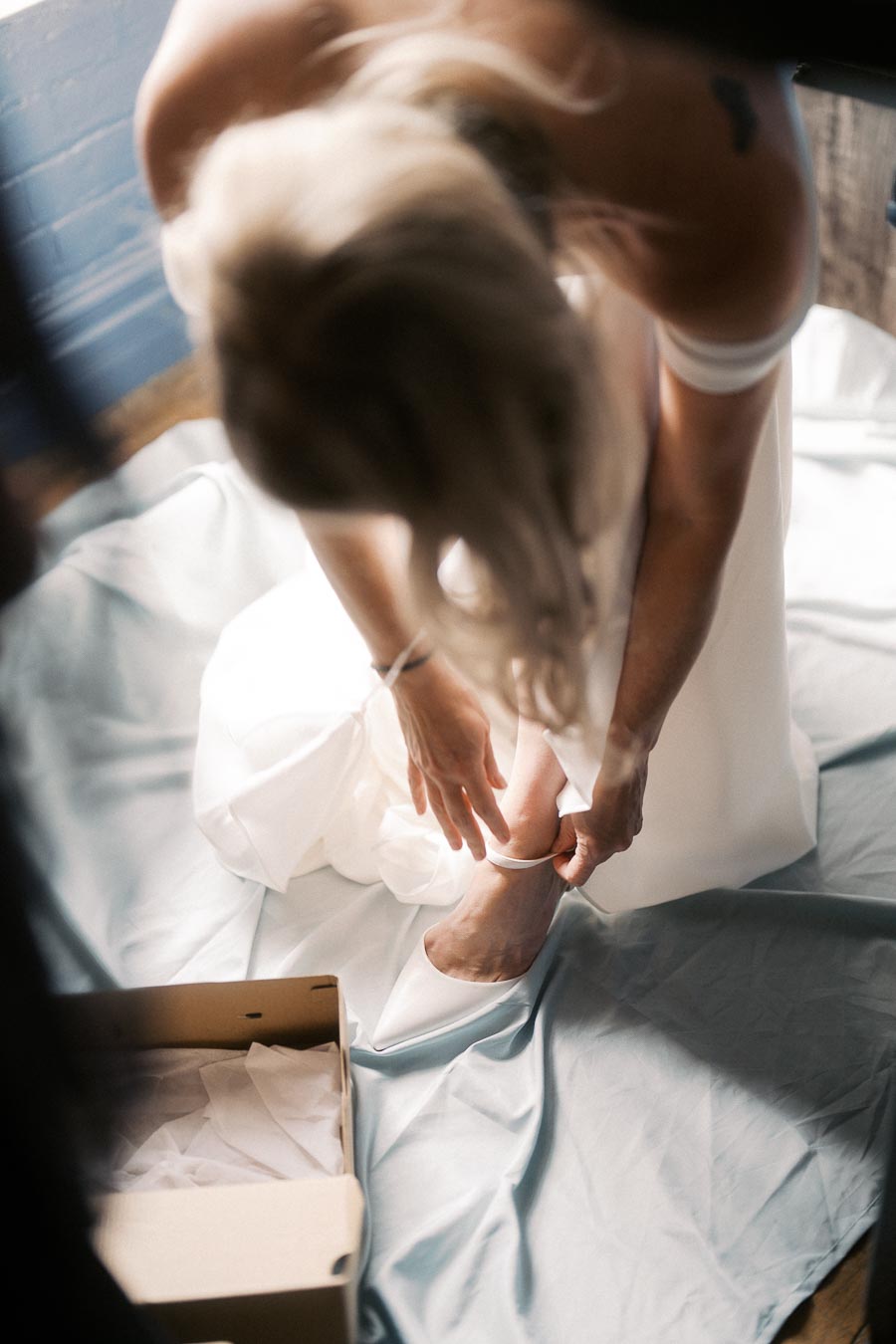 Bride adjusting white shoes on wedding day, wearing off-shoulder dress, sitting on a draped fabric with shoe box nearby.