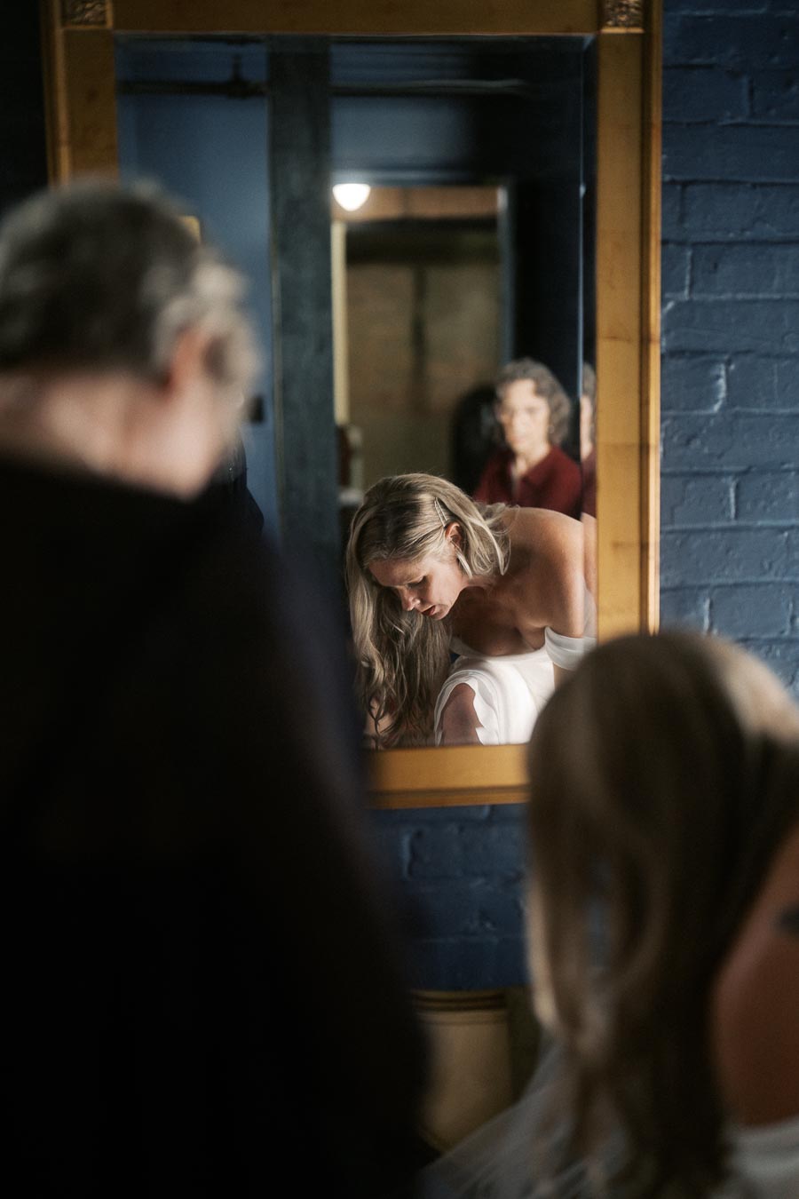 A woman adjusting her outfit in front of a mirror, surrounded by a dark blue brick wall, reflecting a thoughtful moment.