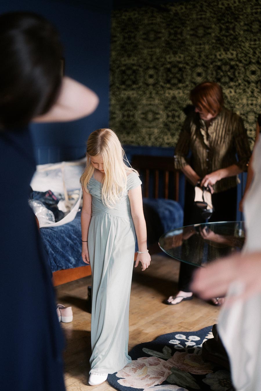 A young girl in an elegant light blue dress stands in a stylish room with a patterned carpet and a decorative wall. She appears contemplative, surrounded by adults adjusting their attire. The setting suggests preparation for a special event or celebration.