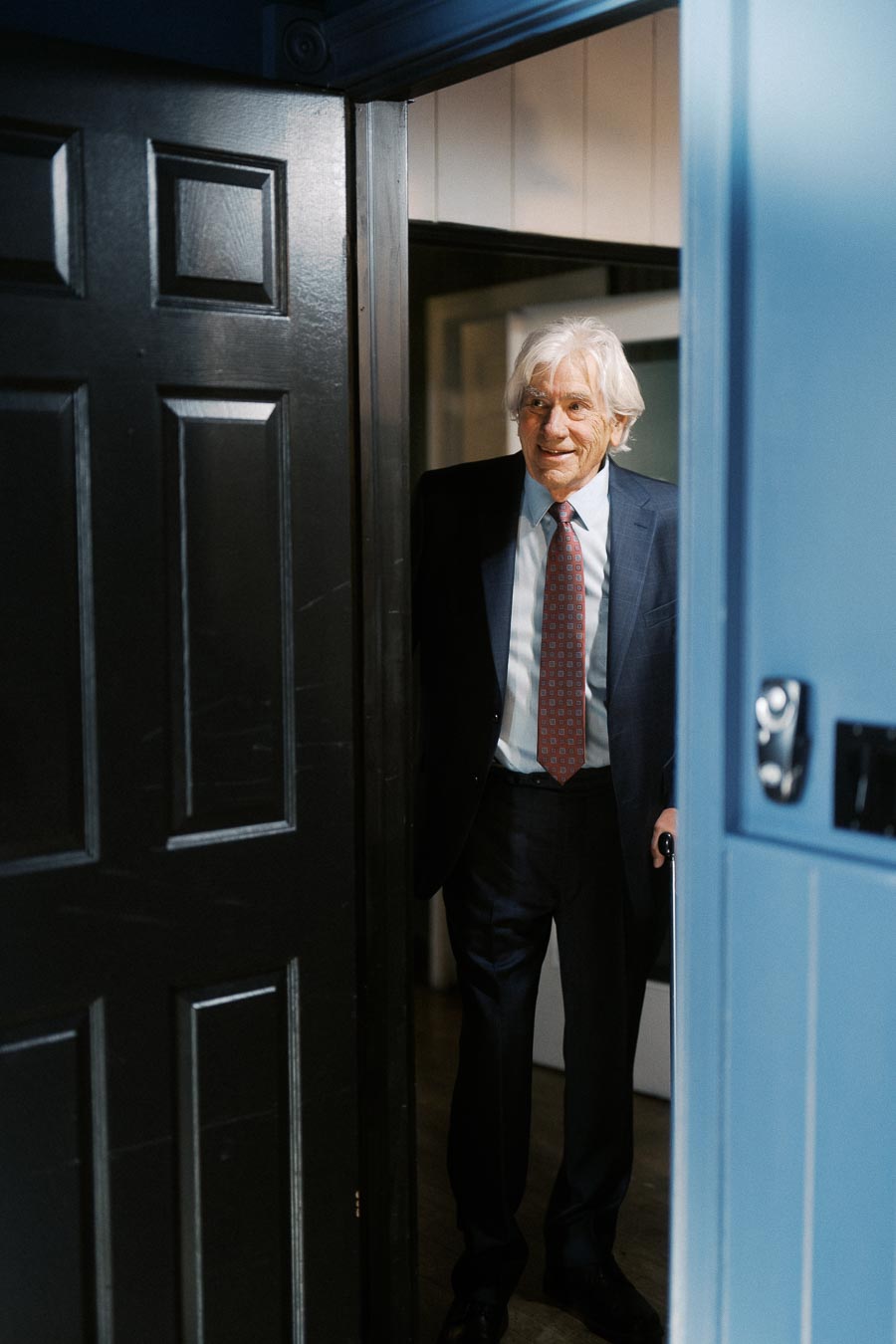 Elderly man in a suit entering a room through a black wooden door, smiling warmly, with bright indoor lighting.