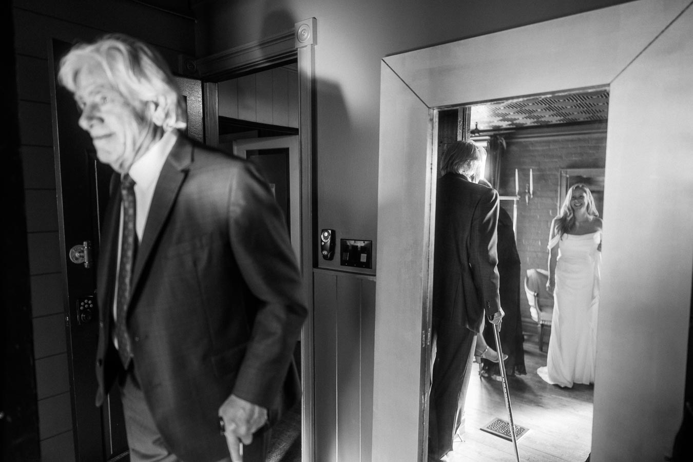 A black and white photograph of a candid moment in a hallway. An older man in a suit, holding a cane, is walking through a door and glancing towards a room where a woman in a wedding dress stands, smiling. The scene captures an intimate and joyous occasion.