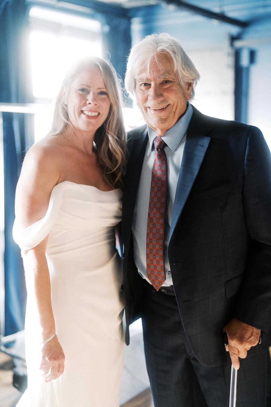 Elderly couple smiling at a formal event, with the woman in a white off-shoulder dress and the man in a dark suit with a red tie, standing together in front of a softly lit window.