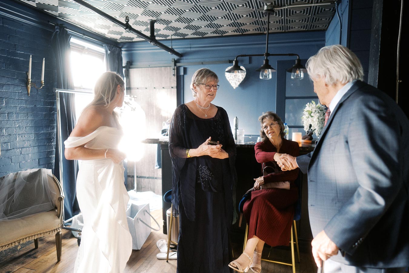 A bride in a white dress interacts with family members in a warmly lit room, featuring elegant decor and a chandelier, creating a joyful atmosphere before a wedding ceremony.