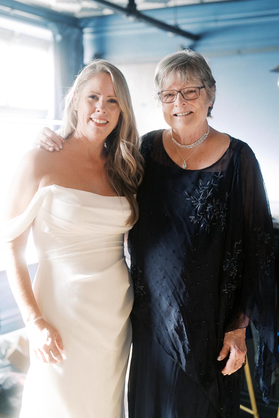Two women smiling together, one in a white wedding dress and the other in a navy blue dress, posing indoors near a window.