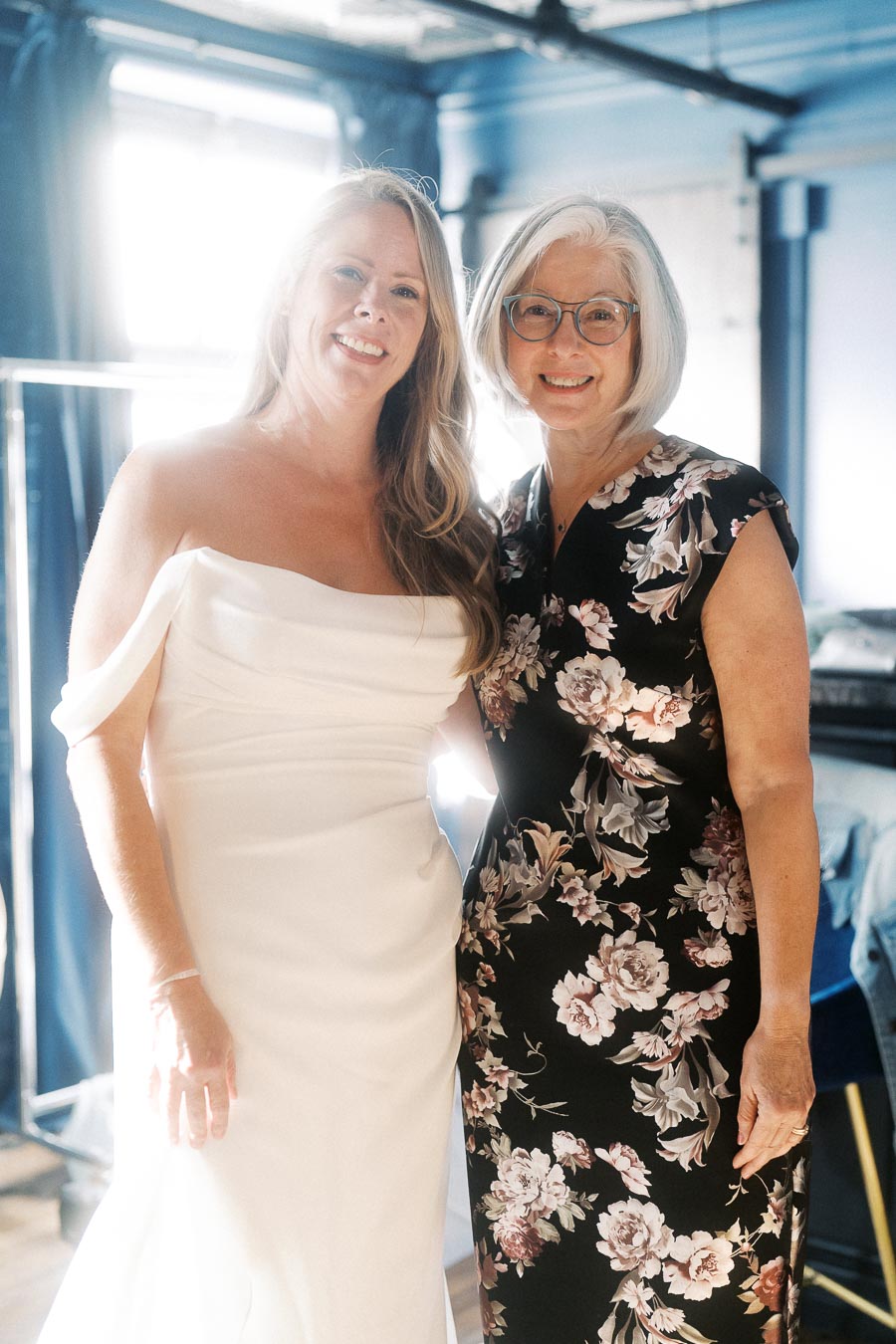 Two women smiling brightly; one in a white off-shoulder wedding gown, the other in a floral-patterned dress, standing in a warmly lit room, suggesting a joyful occasion like a wedding celebration.