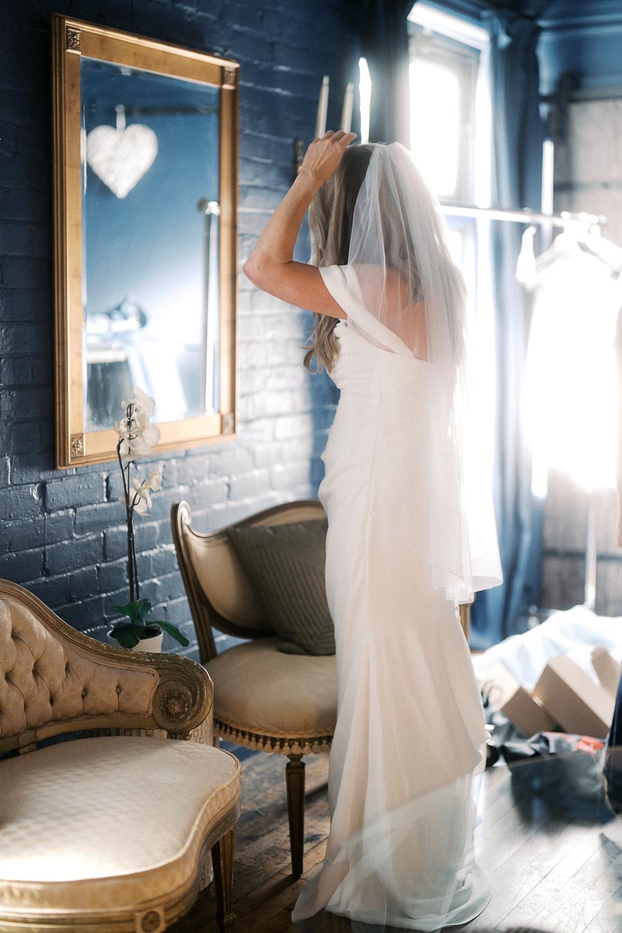 Bride adjusting veil in elegant bridal gown, standing in a vintage-inspired room with a large mirror and soft natural lighting.