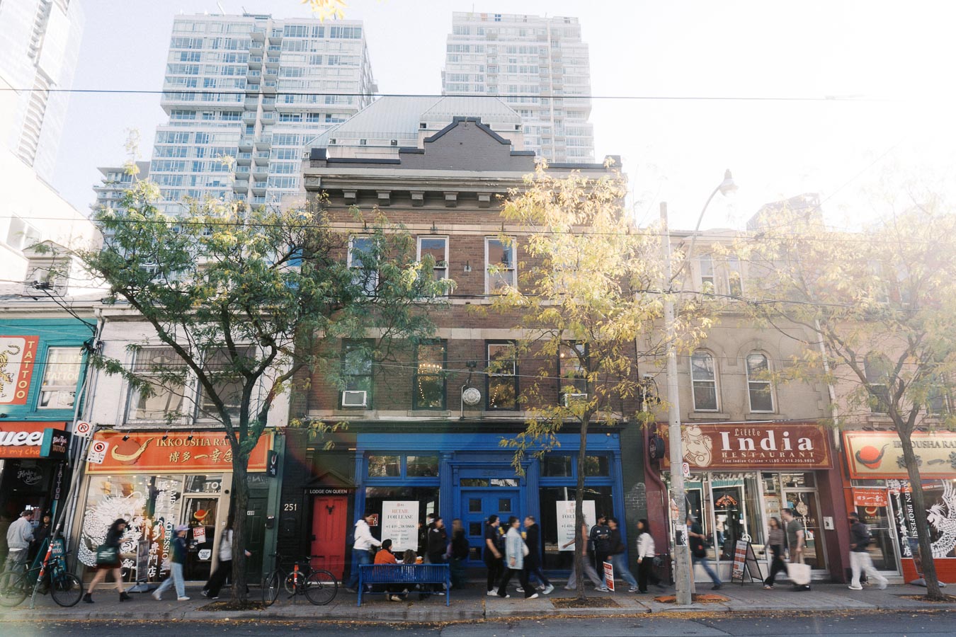 Street view of vibrant urban buildings with shops and restaurants, including a tattoo parlor, ramen shop, and Indian restaurant, lined by trees and pedestrians, against a backdrop of modern high-rise apartments.