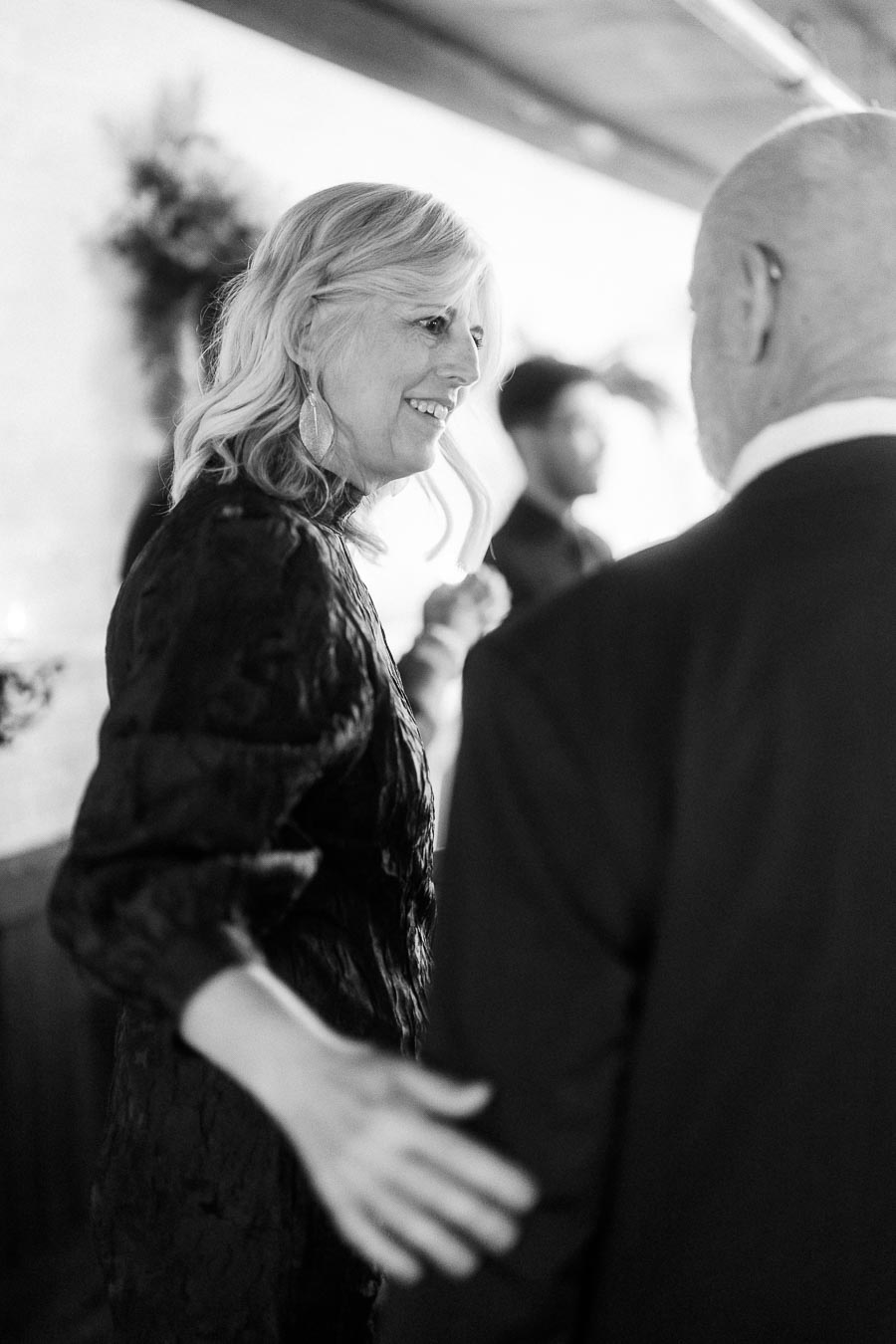 Black and white image of a woman in an elegant dress smiling while talking to a man at a social gathering.