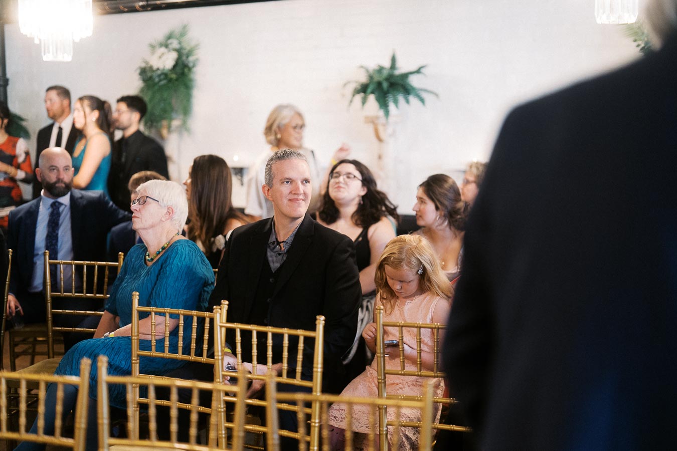 Guests seated at a formal event, featuring a mix of adults and children in elegant attire, while others converse in the background with decorative greenery adorning the space.