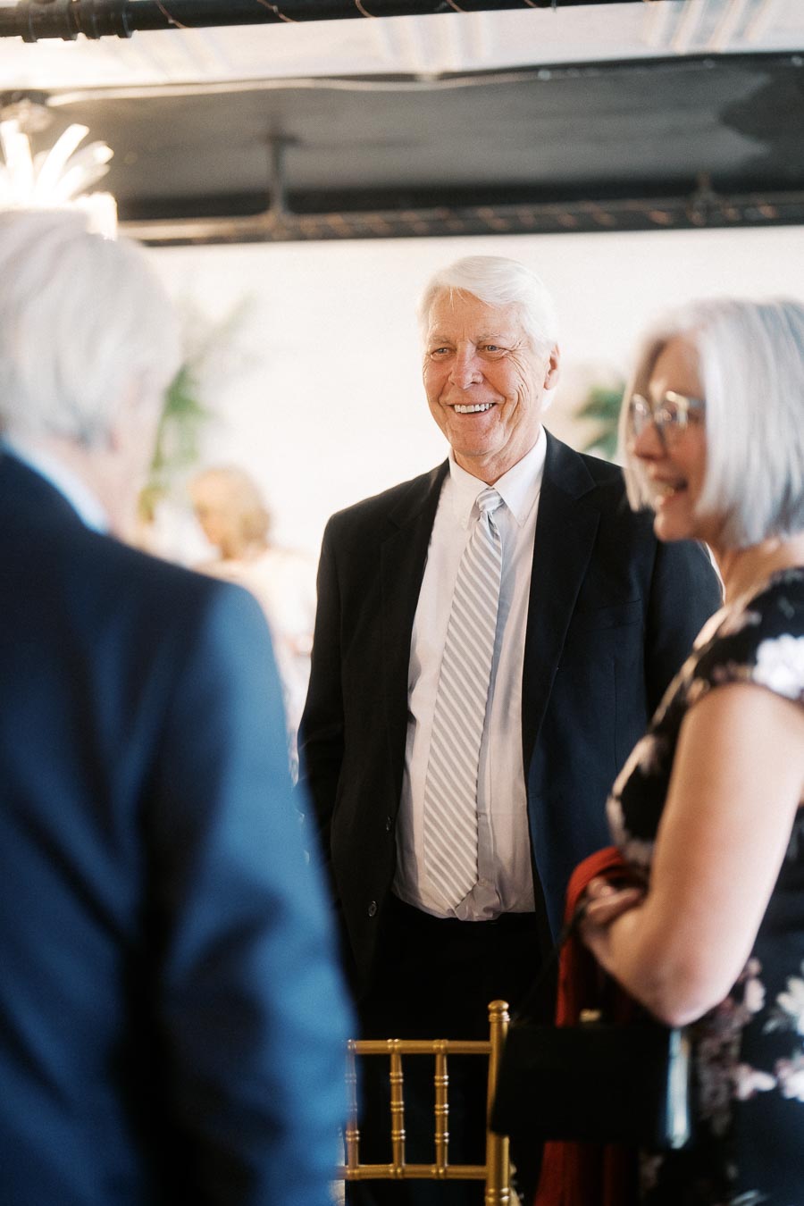 A group of elegantly dressed people engaging in conversation at a formal event, with one man smiling and wearing a suit and tie.