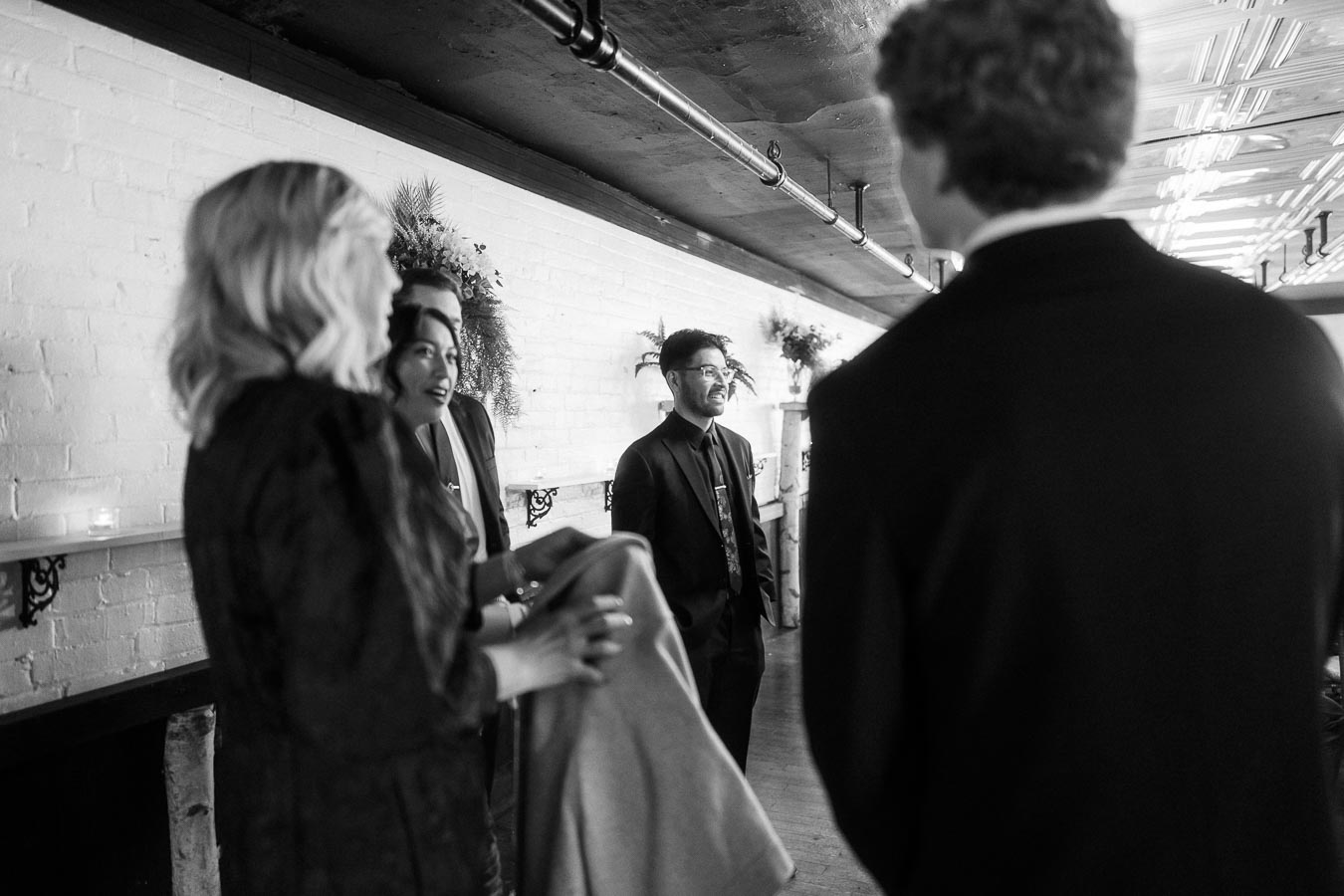 Black and white image of a group of people in formal attire standing and conversing in a stylish indoor setting with decorative plants on a ledge against a white brick wall.