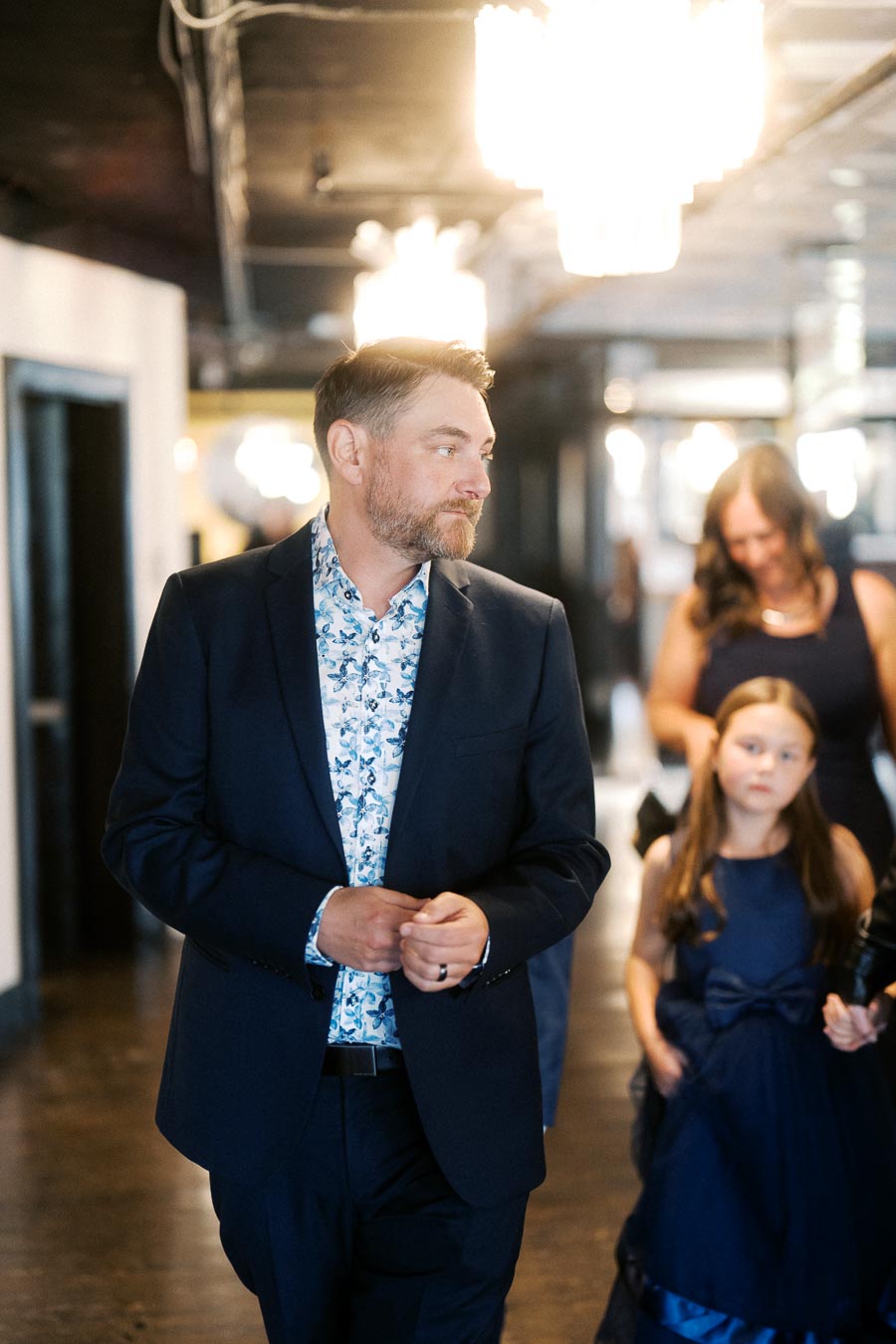 Man in a dark suit and floral shirt walking in a stylish corridor with a woman and a girl in blue dresses in the background. Elegant event setting.