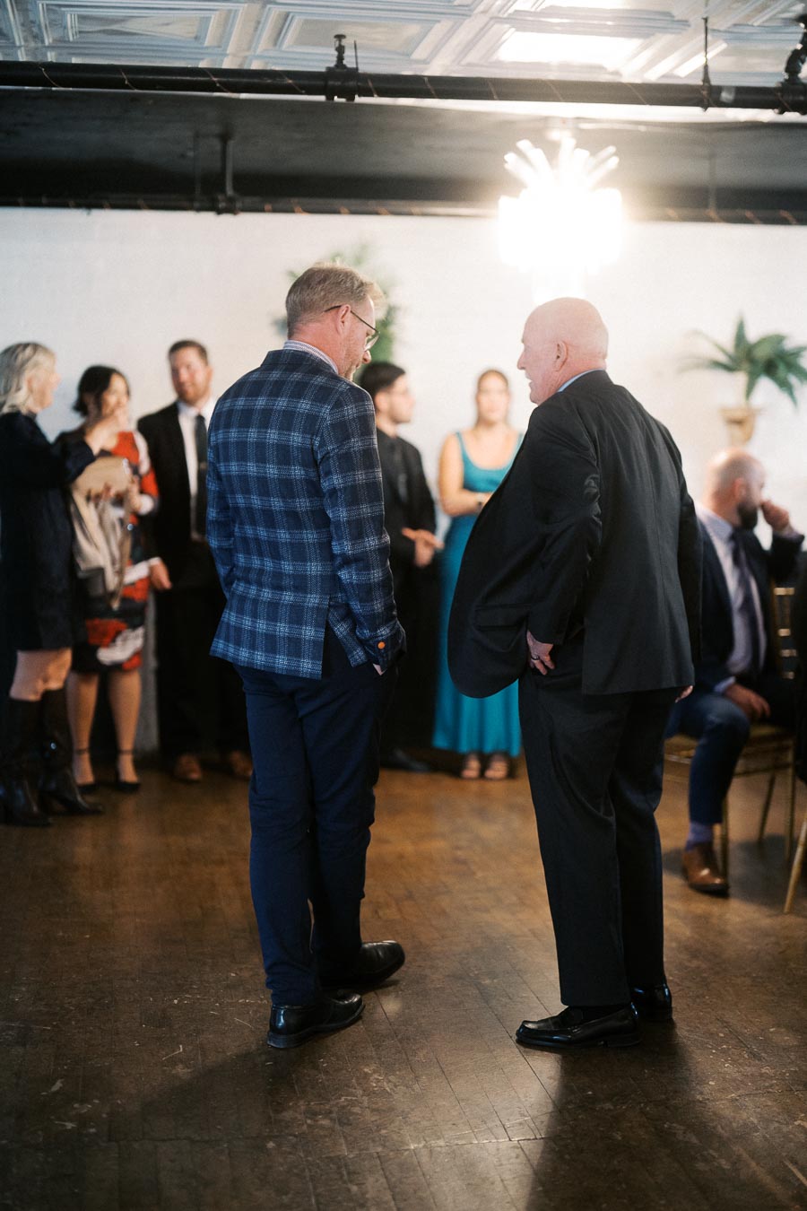 A group of people engaging in conversation at a formal event, with two men in suits as the focal point in the foreground, surrounded by others in elegant attire in a dimly lit room.