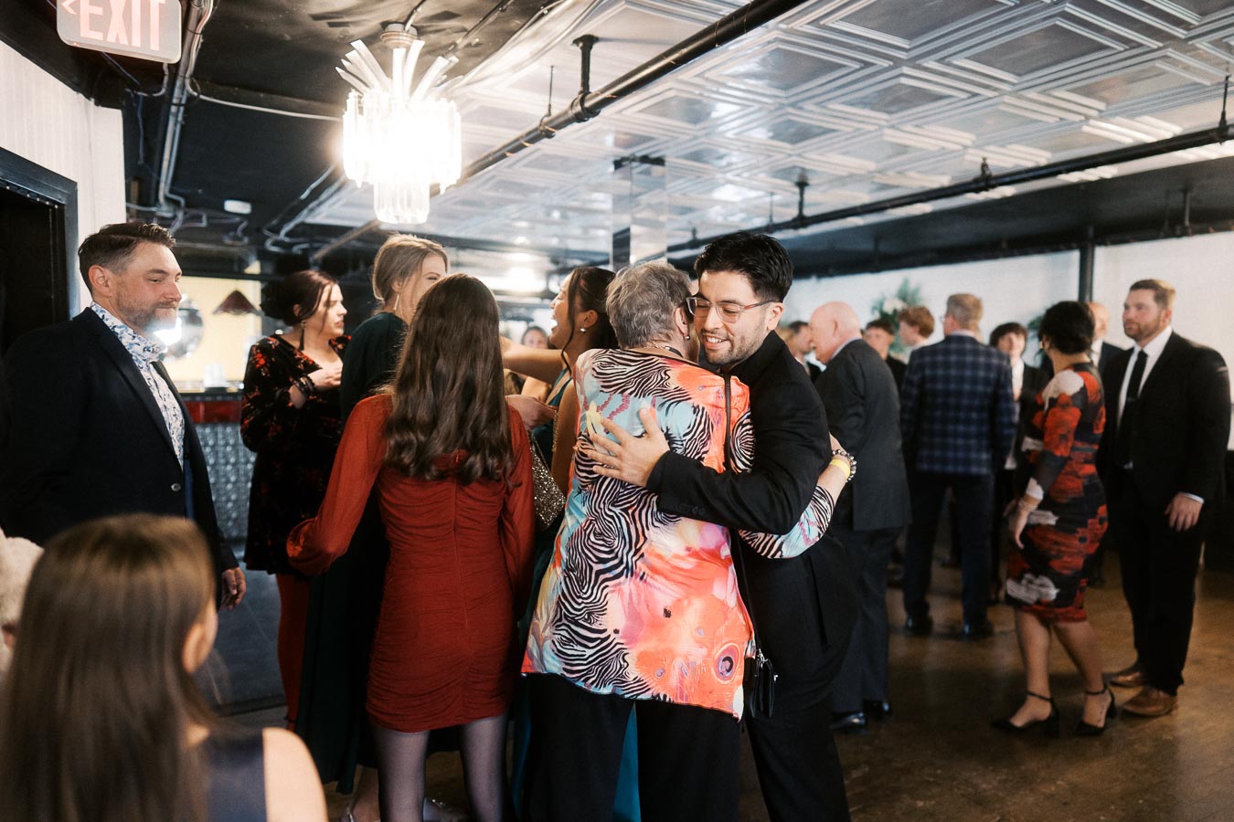 Group of people warmly embracing at a social gathering, with others mingling in a stylish venue under elegant ceiling lights.
