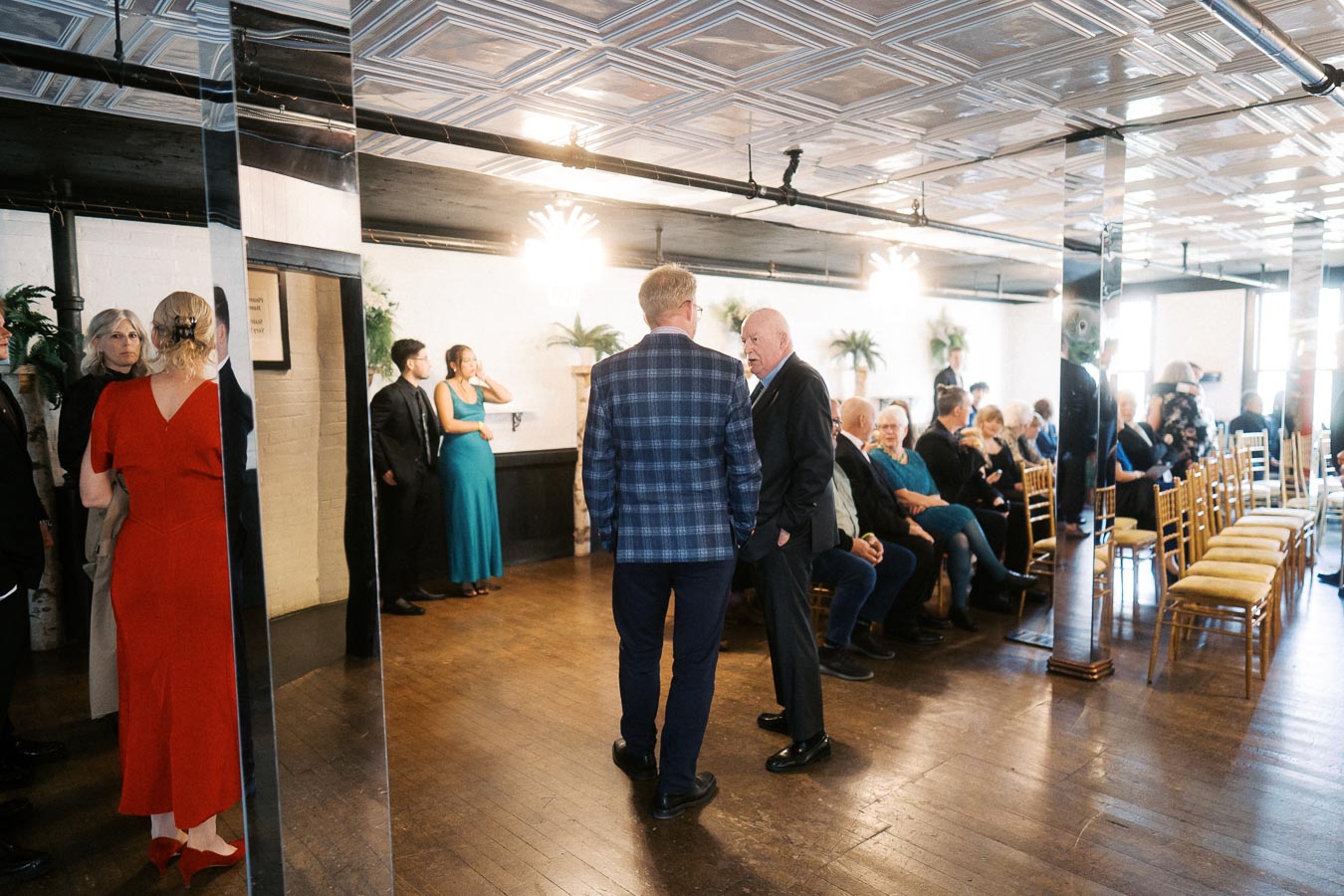 A group of people gathered in a stylish, modern event space with wooden flooring and decorative plants. Guests are engaged in conversation, with some seated on arranged chairs. The setting suggests a formal gathering or reception.