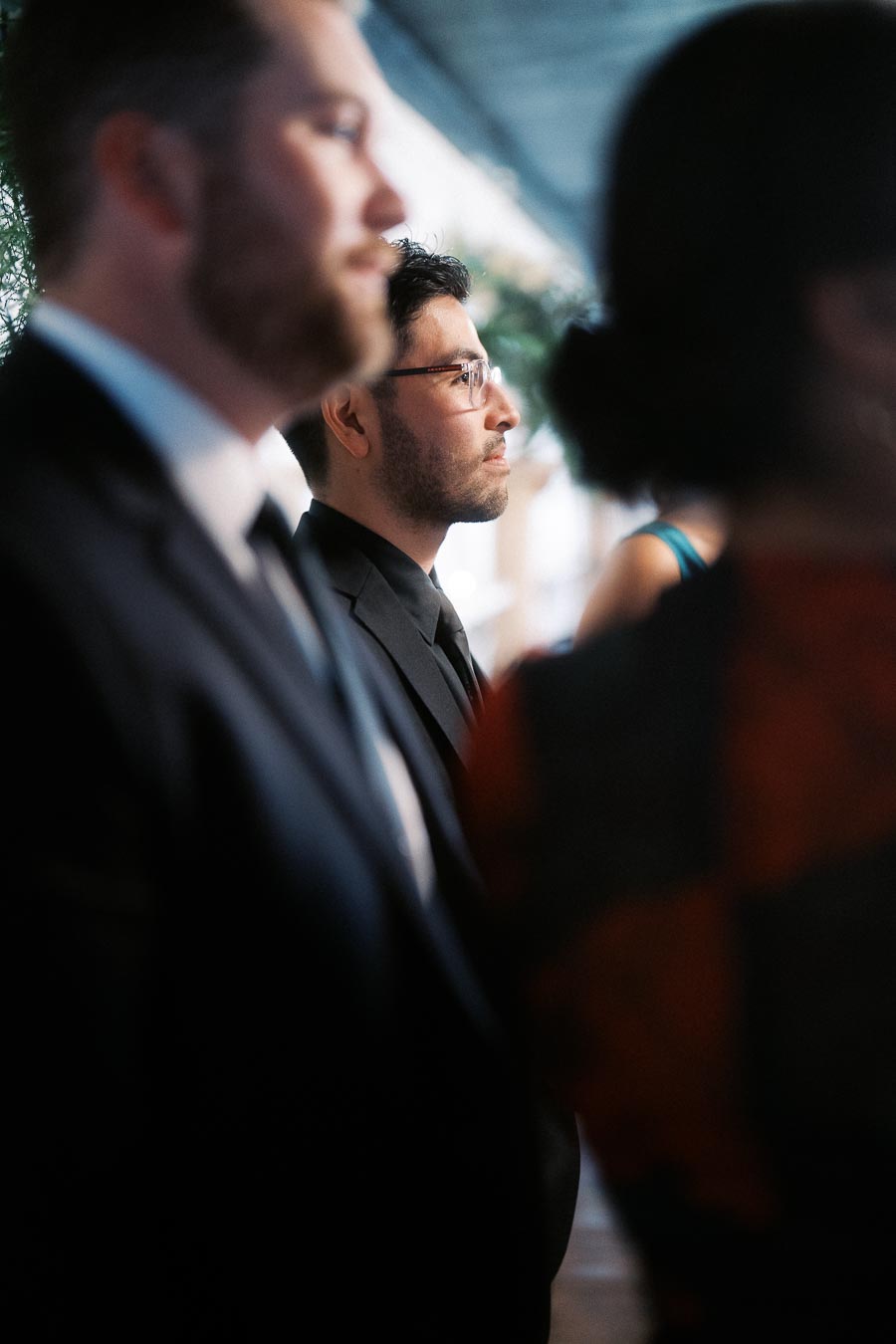 A group of people dressed in formal attire, including suits and ties, attending an indoor event, with a soft focus and natural lighting highlighting the elegant atmosphere.