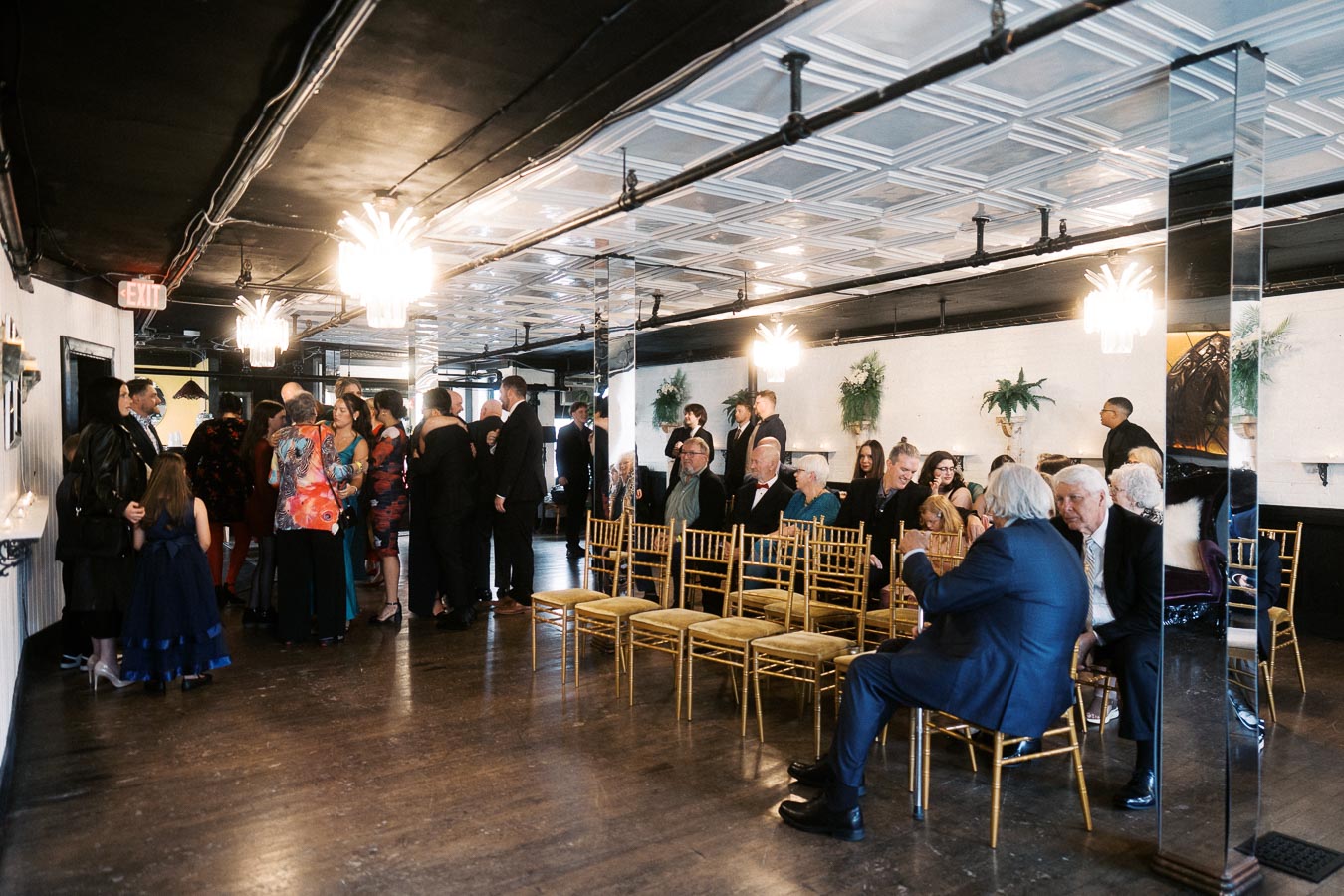 A group of elegantly dressed guests socializing and sitting on gold chairs in a stylish, modern venue with decorative lighting, preparing for an indoor event or ceremony.