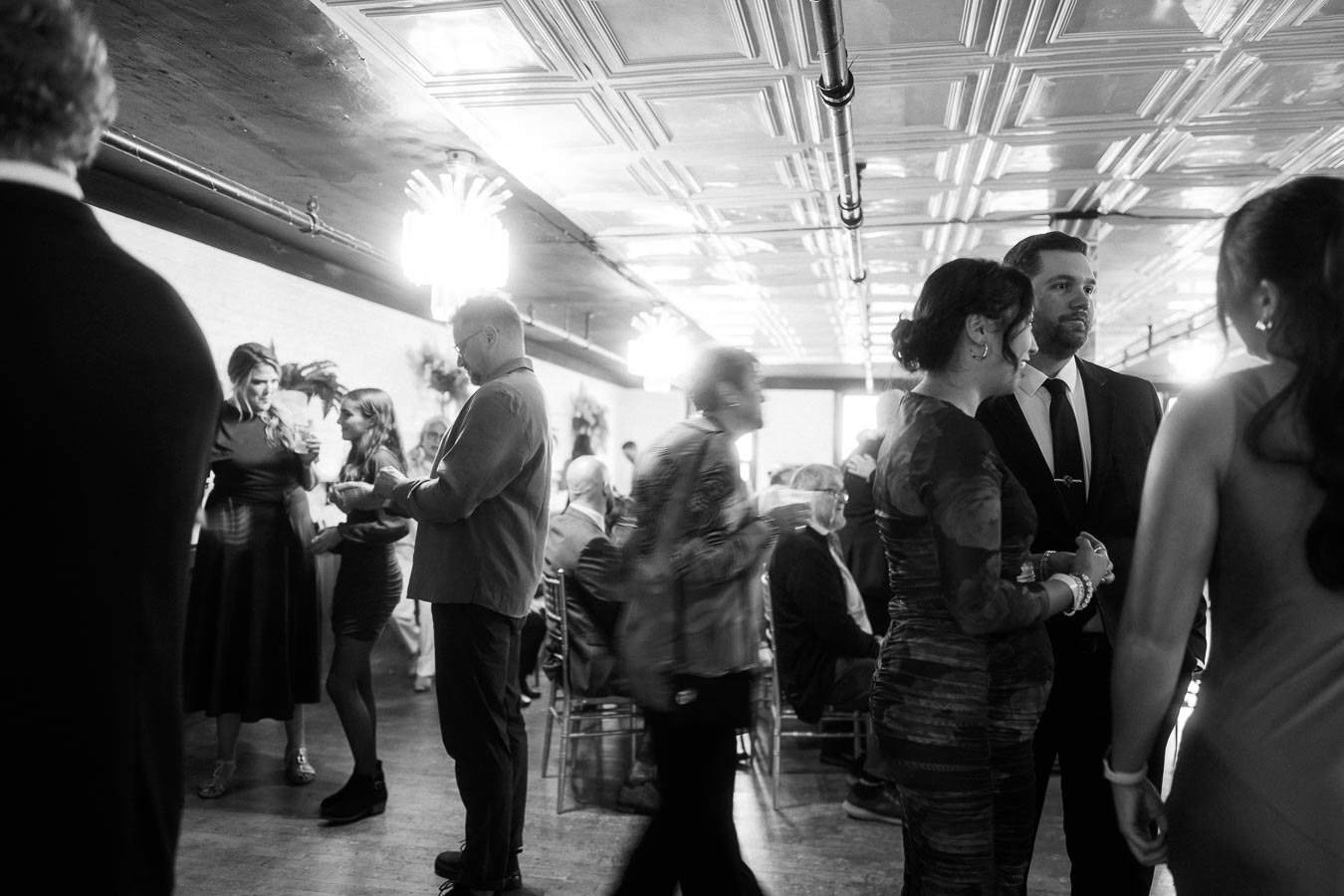 Black and white photo of a lively indoor social gathering, featuring elegantly dressed people engaged in conversations and mingling under decorative ceiling lights.