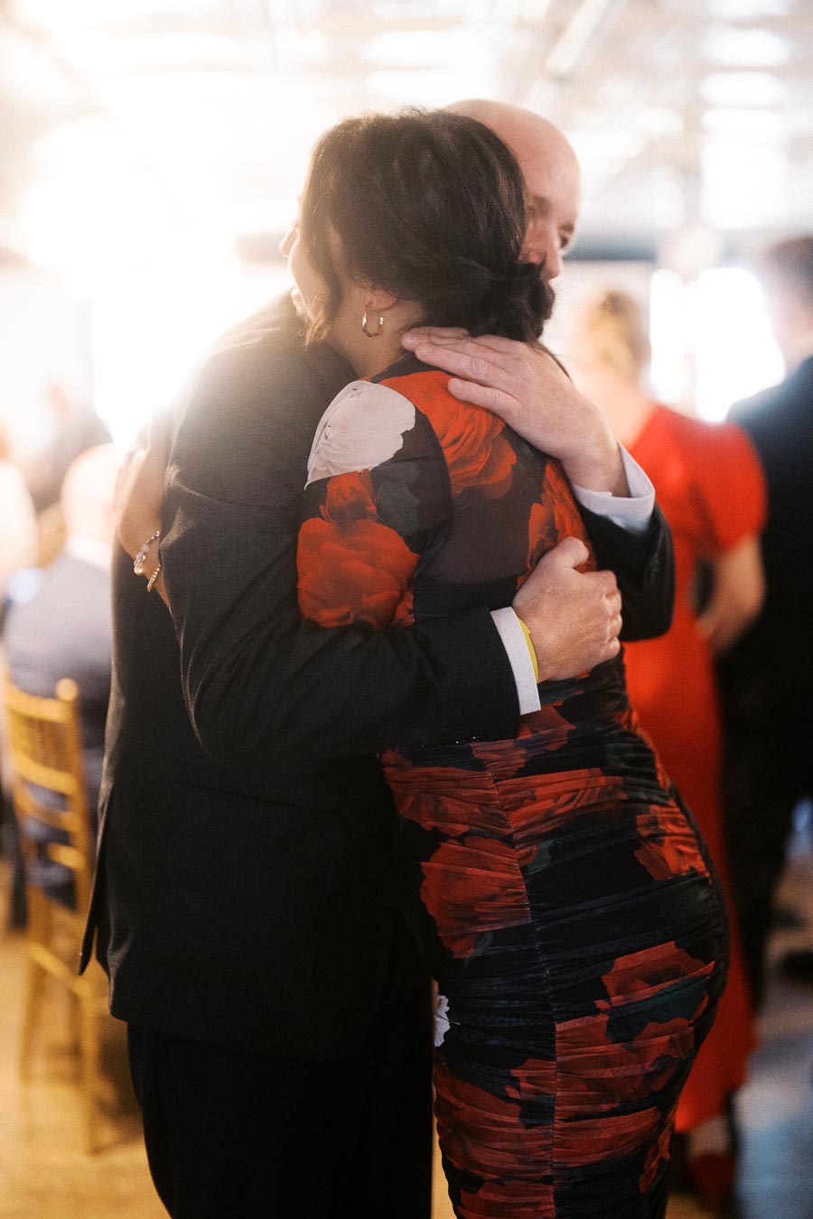A couple warmly embracing at a formal event, with the woman wearing a floral patterned dress and the man in a suit, creating a heartfelt moment filled with emotion and companionship.