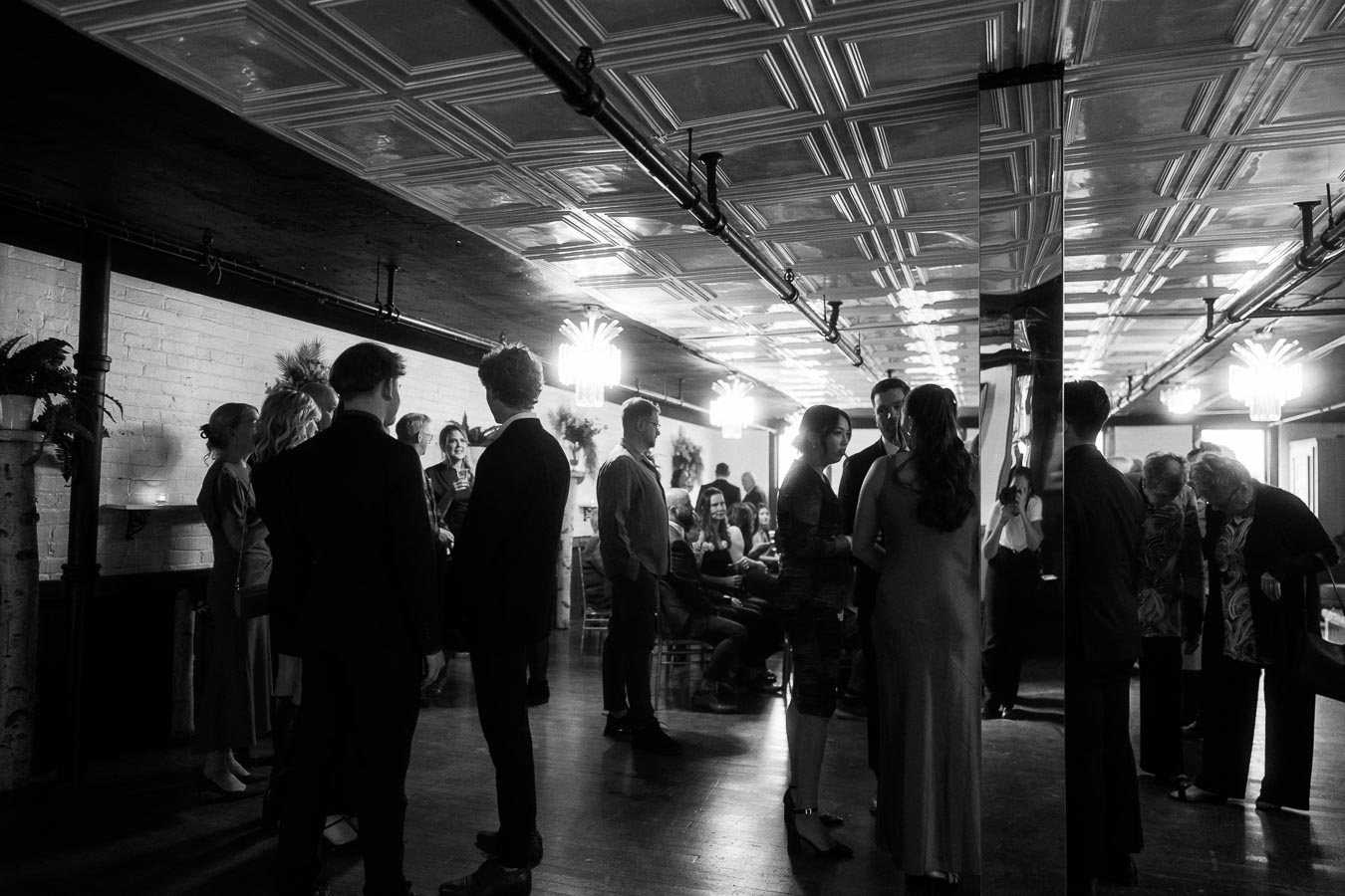 Black and white photo of a social gathering in a stylish venue with decorative ceilings, featuring groups of people engaged in conversation.