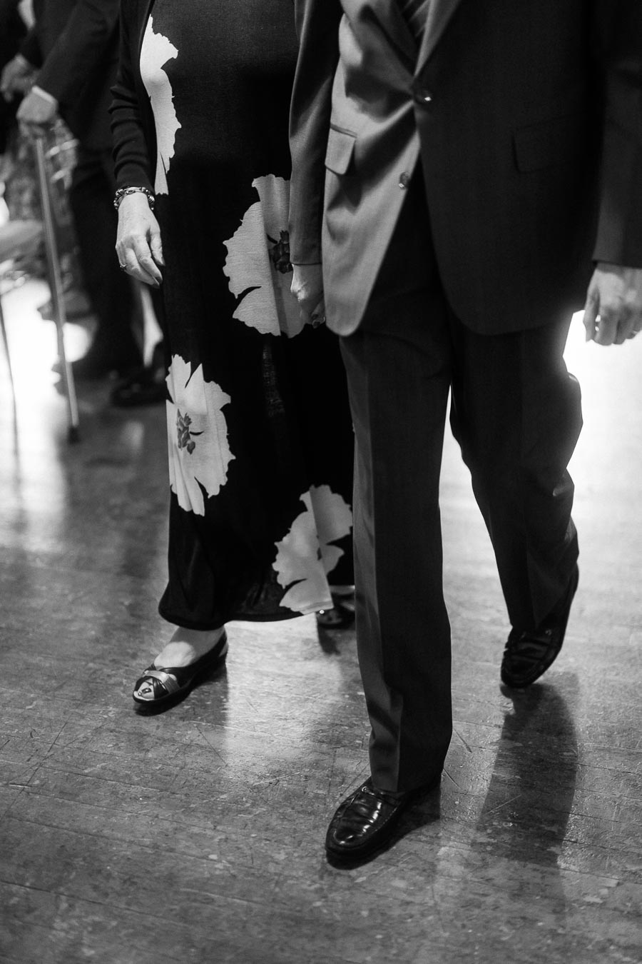 Black and white image of a man in a suit and a woman in a floral dress walking side by side, emphasizing elegance and companionship on a polished floor.