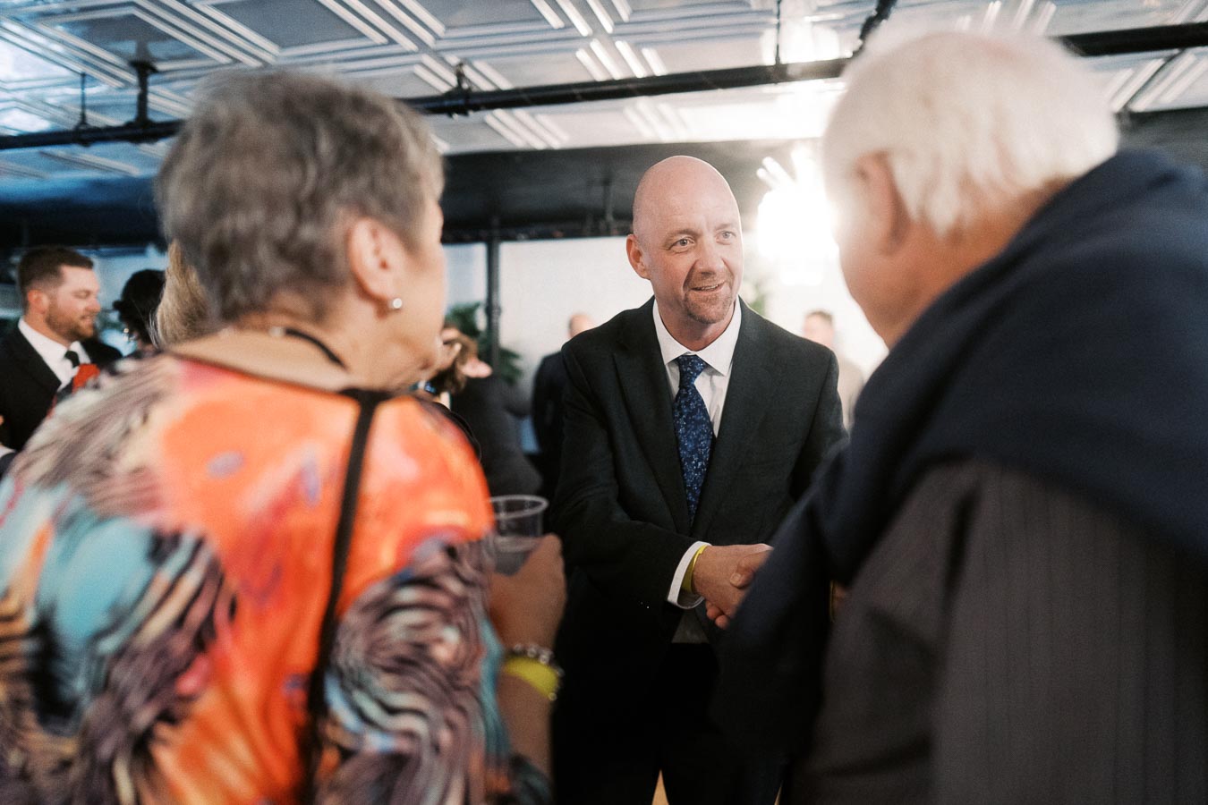 Middle-aged man in a suit shaking hands with another man at a social gathering, surrounded by casually dressed individuals in an indoor setting with modern ceiling tiles.