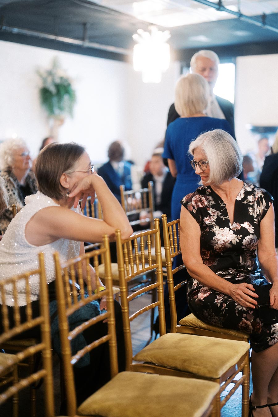 Two women engage in conversation at a formal gathering with golden chairs and elegantly dressed guests in the background.