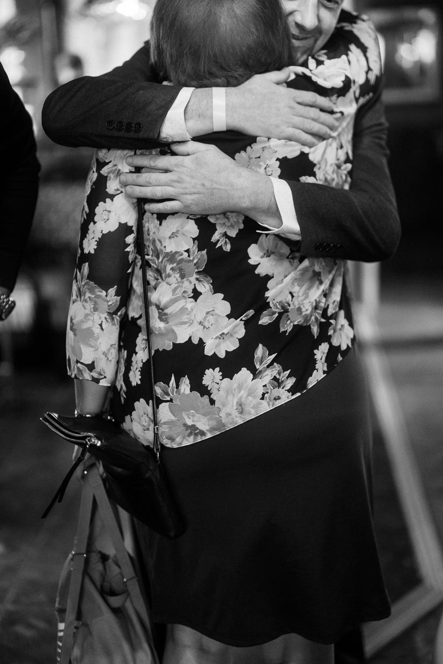A black and white photo capturing a heartfelt hug between two people, one wearing a floral-patterned dress and holding a bag, evoking warmth and connection.