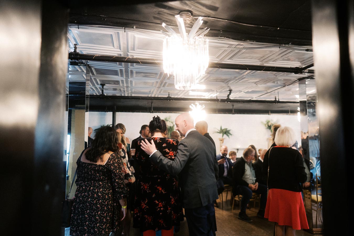 A group of people gather in a warmly lit room with ornate ceiling tiles and a chandelier, enjoying a social event with elegant decor.