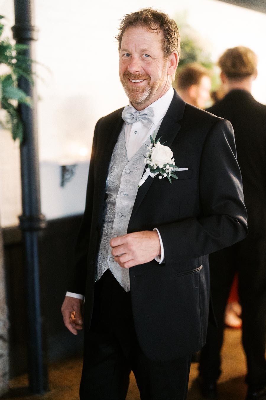 A smiling groom in a formal black tuxedo with a silver vest and white bow tie, adorned with a boutonniere, standing indoors at a wedding venue.