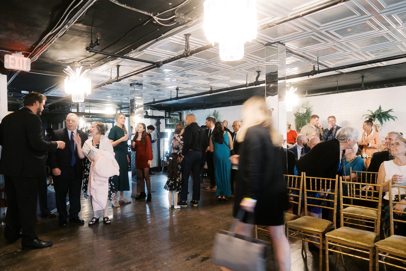 Group of people socializing at an indoor event in a well-lit venue with chandeliers and wooden flooring, filled with lively conversation and interactions.